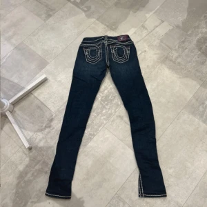 True Religion jeans med kontrastsömmar - Snygga blå jeans från True Religion med tydliga vita kontrastsömmar och ikoniska bakfickor. Lila patch bak i midjan och detaljer i lila vid fickorna. Modellen är slim och har låg midja. Perfekta för dig som vill sticka ut med klassisk streetstyle.