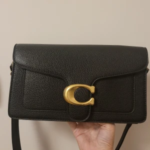 Svart axelväska från Coach i skinn - Snygg svart axelväska från Coach i äkta skinn med en stor guldfärgad C-detalj på framsidan. Stilren och klassisk design med justerbar axelrem. Perfekt för dig som vill ha en tidlös och trendig accessoar med lyxig känsla.