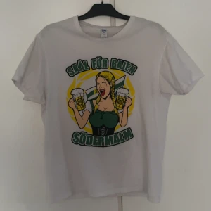 Hammarby tröja - Vit t-shirt från JHK i 100% bomull med tryck framtill: en tecknad tjej med flätor, två ölglas och texten 'Skål för Bajen Södermalm' i grönt och gult. Klassisk passform och rund hals. Perfekt för dig som vill visa ditt stöd för Bajen.