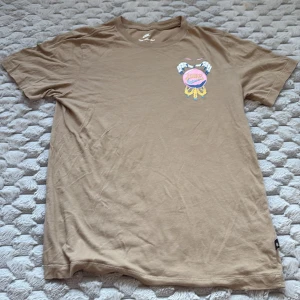 Beige Nike t-shirt med tryck - Säljer en beige t-shirt från Nike i bomull med coolt färgglatt tryck på både bröstet och ryggen. T-shirten har klassisk passform och korta ärmar. Perfekt för dig som gillar streetstyle och snygga prints.
