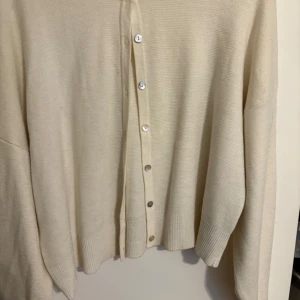 Beige kofta med knappar från Gina Tricot - En stilren beige kofta med långa ärmar och knappar framtill. Koftan är stickad i ett mjukt material och har ribbade muddar vid ärmslut och nederkant. Perfekt för lager-på-lager och enkel att matcha med olika outfits. Använd fåtal gånger