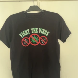 Hammarby tröja - Cool svart t-shirt med färgglatt tryck på bröstet där det står 'FIGHT THE VIRUS' och tre symboler med röd cirkel och streck. Klassisk rund hals och korta ärmar. Perfekt statement-plagg för dig som vill sticka ut och visa attityd. Trycket är lite trasigt 