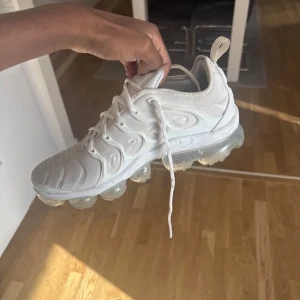 Nike Air Vapormax Plus vita sneakers - Nike Air Vapormax Plus i helvitt med genomskinlig sula och snygga detaljer på ovandelen. Skorna har snörning och en futuristisk design med vågiga linjer och meshmaterial. Perfekta för dig som vill sticka ut med en modern och sportig look.