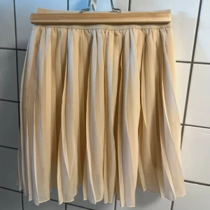 Plisserad beige kortkjol - Säljer en superfin plisserad kjol i ljus beige ton. Kjolen är kort och har ett luftigt, flowigt tyg som ger snygg volym. Använd 2-3 gånger