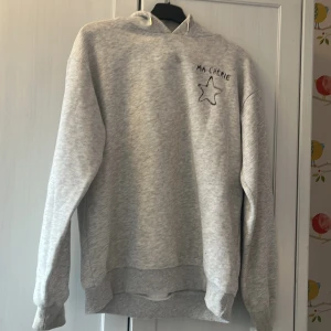Grå hoodie med tryck 'Ma Chérie' och stjärna - Säljer en ljusgrå hoodie med svart tryck där det står 'Ma Chérie' och en stjärna på bröstet. Tröjan har huva, långa ärmar och ribbade muddar. Perfekt för dig som gillar en chill och enkel stil. Finns några fläckar som kanske går bort i tvätten!