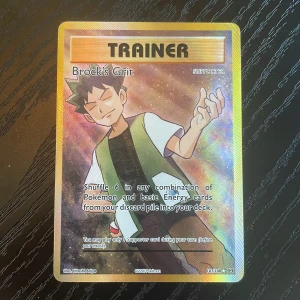 Brock's Grit - Cool och glittrig Pokémon-tränarkort med Brock! Perfekt för dig som vill stärka din lek och samla på unika supporter-kort. Passar både samlare och spelare som gillar Pokémon-världen.