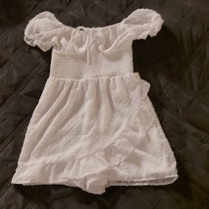 Vit kort offshoulder klänning - Supersöt vit kort one- piece med offshoulder puffärmar och smockad överdel. Shortsen har volangkant och tyget är lätt med små prickar i strukturen. Perfekt för sommarens alla tillfällen och ger en romantisk vibe.