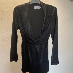 Svart omlottklänning NA-KD x Hannalicious - Supersnygg svart kort omlottklänning från NA-KD x Hannalicious. Klänningen har lång ärm, djup v-ringning och markerad. Materialet är glansigt och känns lyxigt, perfekt för att sticka ut. Enkel men ändå elegant design. Saknar bältet men går att ha det utan eller med ett annat svart bälte till.