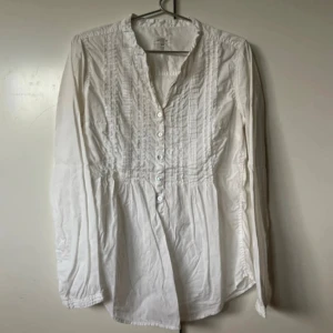 Vit henley-skjorta med knappar - Vit henley-skjorta från H&M med lång ärm och dekorativa veck framtill. Skjortan har flera knappar längs bröstet och är tillverkad i mjuk bomull. Perfekt för en avslappnad men ändå stilren look.