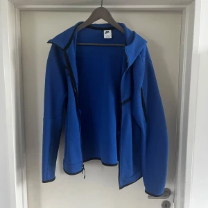 Blå Nike vindjacka med huva - (Byxorna finns också) Blå vindjacka från Nike med svart logga på bröstet, hel dragkedja och huva. Jackan har svarta detaljer vid dragkedjor och muddar samt en extra ficka med dragkedja på ärmen. Perfekt för dig som gillar sportig och clean stil.