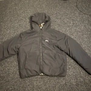 Reversibel teddyjacka från Stüssy - Säljer en fet reversibel teddyjacka från Stüssy med huva. Ena sidan är svart med diskret logga och dragkedja, andra sidan är fluffig med mönster i vitt, svart och brunt samt bröstficka med logga. Perfekt för dig som vill switcha stil snabbt.