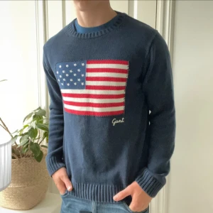 Mörkblå stickad tröja från Gant - Säljer en mörkblå stickad tröja från Gant med en stor amerikansk flagga på bröstet. Tröjan har rund halsringning, ribbade muddar och är långärmad. Perfekt för dig som gillar klassisk preppy stil och vill sticka ut lite extra.