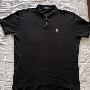 Svart piké från Polo Ralph Lauren - Svart pikétröja från Polo Ralph Lauren i storlek L. Klassisk design med krage, korta ärmar och två knappar framtill. Ikonisk vit broderad logga på bröstet. Mjuk och skön bomull som passar perfekt till jeans eller shorts.