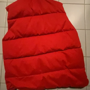 Röd dunväst från Canada Goose - Säljer en klassisk röd dunväst från Canada Goose med hög krage och breda, quiltade paneler. Västen har tryckknappar framtill och en diskret logga på bröstet samt en patch vid ena fickan. Perfekt för lager-på-lager och riktigt snygg till streetwear.