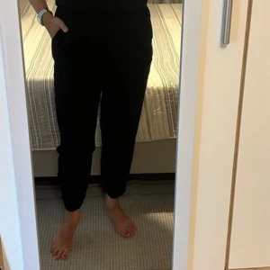 Svart omlott byxdress med resår - Snygg svart byxdress med omlott överdel och resår i midjan. Byxbenen är avsmalnande och avslutas med resår vid anklarna. Ärmlös modell i mjukt tyg, perfekt för en stilren look. Enkel att matcha med sneakers eller klackar.