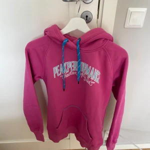 Rosa hoodie från Peak Performance M - Snygg rosa hoodie från Peak Performance med vit och blå logga framtill. Tröjan har huva med blå snören och en stor magficka. Perfekt för dig som gillar sportig och avslappnad stil. Mjuk insida och långärmad modell.