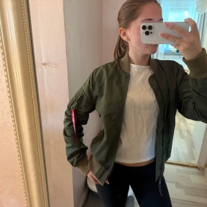 Grön bomberjacka från Alpha Industries - Säljer en klassisk grön bomberjacka från Alpha Industries med orange foder och ikonisk röd tag på ärmen. Jackan har ribbade muddar, dragkedja framtill och fickor på ärmen. Perfekt för dig som vill ha en streetig och tidlös look.