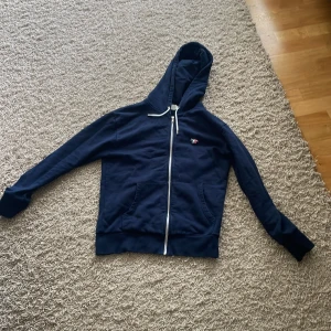 Marinblå hoodie från Maison Kitsuné - Snygg marinblå hoodie från Maison Kitsuné med dragkedja och huva. Tröjan har vita snören, två fickor fram och en broderad rävlogga i blått, vitt och rött på bröstet. Perfekt för dig som gillar stilrena och bekväma plagg.