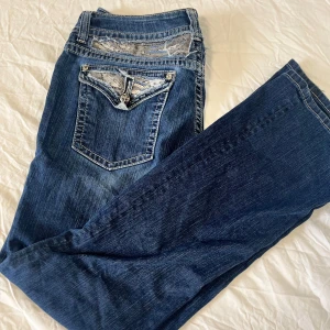 Miss Me jeans med paljettdetaljer - Snygga blå jeans från Miss Me med unika paljettdetaljer och spets på bakfickorna. Jeansen har kontrastsömmar, dekorativa knappar och klassisk femficksdesign. Perfekta för dig som vill sticka ut med lite extra bling i vardagen.