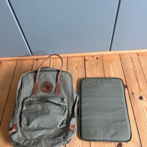  Grön Fjällräven Kånken ryggsäck - Säljer en klassisk grön Fjällräven Kånken ryggsäck med bruna skinnhandtag och detaljer. Ryggsäcken har en stor ficka framtill med dragkedja, justerbara axelband och medföljande datorfodral. Perfekt för skola eller cityhäng.