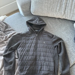 Svart cardigan från Polo Ralph Lauren - Säljer en svart hoodiejacka från Polo Ralph Lauren med quiltad front och ärmar i mjuk bomullsmix. Jackan har huva med snörning, dragkedja framtill och broderad logga på bröstet. Perfekt för dig som gillar stilren och snygg look.