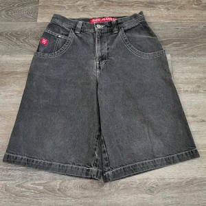 JNCO Jeans svarta loose shorts drake - Grymma svarta jeansshorts från JNCO Jeans med bred passform och hög midja. Baksidan har en stor röd broderad drake på fickan och coola kontrastsömmar. Klassisk femficksdesign och tydliga JNCO-loggor. Perfekt för dig som gillar streetwear och vill sticka ut.