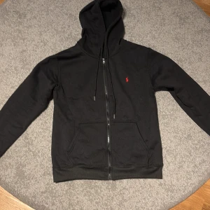 Svart hoodie från Ralph Lauren - Svart hoodie från Ralph Lauren med dragkedja och huva. Klassisk röd broderad logga på bröstet, kängurufickor och snörning i huvan. Tillverkad i mjuk bomull, perfekt för chill dagar. Enkel och stilren design som funkar året runt.