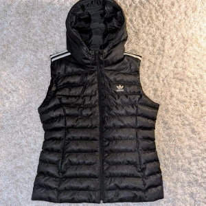 Svart väst med huva från Adidas - Svart dunväst från Adidas med huva och dragkedja framtill. Västen har de klassiska vita ränderna på axlarna och Adidas-logga på bröstet. Quiltad design med horisontella sömmar och diskret mönster i tyget. Perfekt för lager-på-lager och streetstyle.