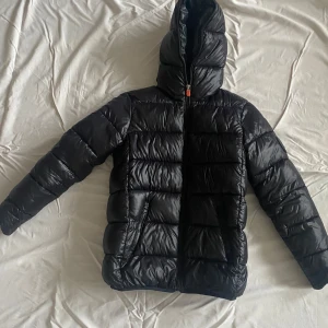 Svart pufferjacka från Save The Duck - Säljer en svart pufferjacka från Save The Duck med huva och dragkedja. Jackan är quiltad och har orange foder med texten '100% Animal Free'. Märke på ärmen. Perfekt för kalla dagar och riktigt snygg streetstil. Storlek xs 164