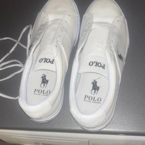 Vita sneakers från Polo Ralph Lauren - Snygga vita sneakers från Polo Ralph Lauren med klassisk logga på sidan och innersulan. Skorna har rund tå, platt sula och snörning. Tillverkade i textilmaterial med vita detaljer och stilren design som passar till det mesta. Storleken är 41 och har använda 2-3 gånger och bara suttit på hyllan ganska länge. De är i bra skick, lita smutsig där framme men inga hål eller något sånt. Priset är ej hugget 