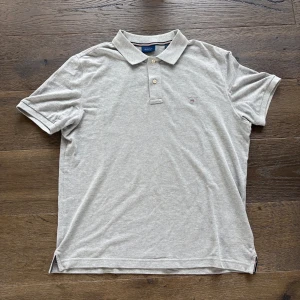 Grå pikétröja från GANT - Snygg grå pikétröja från GANT med klassisk krage och två knappar framtill. Tröjan har korta ärmar och en liten broderad logga på bröstet. Perfekt för en chill och stilren look. Materialet är mjuk bomull som känns skönt mot huden. Bara att fråga på vid funderingar 👍
