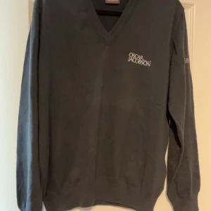 Mörkgrå v-ringad tröja Oscar Jacobson - Snygg mörkgrå v-ringad tröja från Oscar Jacobson med diskret logga broderad på bröstet och ärmen. Tröjan har långa ärmar och är gjord i mjuk bomull, perfekt för dig som gillar stilrena och klassiska plagg. Passar dig som vill ha en clean look.