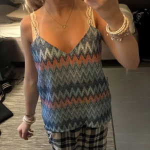 Mönstrad linnetopp - Säljer en snygg linnetopp med färgglatt zickzackmönster i blått, rosa och vitt. Toppen har smala axelband med spetsdetaljer och en v-ringad halsringning. Perfekt för varma dagar och ger en cool, avslappnad vibe.