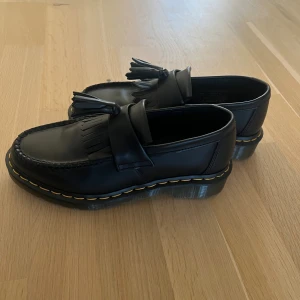 Dr. Martens Adrian loafers - Klassiska svarta Dr. Martens Adrian loafers i skinn med tofsar och fransdetalj framtill. Grov sula med den ikoniska gula sömmen runt om. Köpta i Italien men i fel storlek så de är inte använda.   Strl 38.