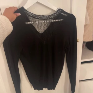 Svart tröja med spetsdetalj - Snygg svart långärmad tröja med v-ringning och transparent spets upptill. Mycket bra skick, säljer då den inte längre används.
