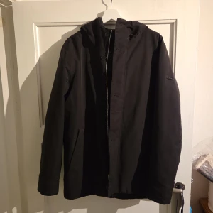 Svart parkajacka från Elvine, M - Säljer en svart parkajacka från Elvine i storlek M. Jackan har huva, dragkedja och knappar framtill samt två fickor med dragkedja. Enkel och stilren design som passar perfekt till kyligare dagar. Materialet känns slitstarkt och jackan har en klassisk passform.