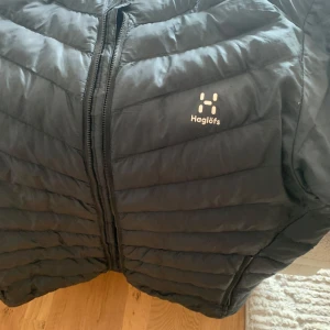 Svart pufferjacka från Haglöfs, L - Säljer en svart pufferjacka från Haglöfs i storlek L. Jackan har en klassisk quiltad design med dragkedja framtill och diskret Haglöfs-logga på bröstet. Perfekt för kyliga dagar och enkel att matcha med olika outfits.