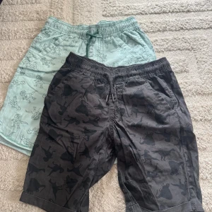 Två mönstrade shorts från Lupilu - Säljer två par shorts från Lupilu, ena i ljusgrön med coolt geometriskt djurmönster och andra i mörkgrått med svarta dinosauriesilhuetter. Båda har elastisk midja och snörning framtill, tillverkade i mjuk bomull. Perfekta för sommaren och chill dagar. Storlek 122/128.