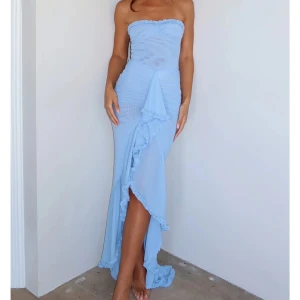 Ljusblå axelbandslös långklänning - Superfin ljusblå långklänning med axelbandslös design och volangdetaljer längs byst och slits. Klänningen har figurnära passform, rynkade partier och en snygg slits framtill. Perfekt för dig som vill sticka ut med en romantisk och trendig look.