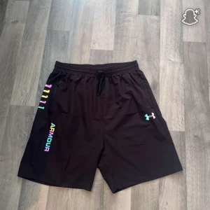 Svarta Under Armour träningsshorts - Svarta träningsshorts från Under Armour med elastisk midja och snörning. Snyggt regnbågsfärgat logotryck på ena benet och klassisk UA-logga på andra. Lätta och luftiga i syntetmaterial, perfekta för gym eller löpning. Den har lite hål ot sidan och en liten hål ner som är jätte small jätte de syns ej jag vistte ej de fanns men de drf de är billig 