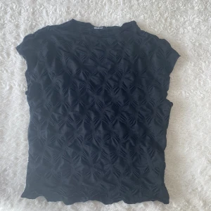 Svart strukturerad topp från Ginatricot - Svart topp från Ginatricot med coolt strukturerat mönster som ger en unik 3D-effekt. Modellen är ärmlös med rund hals och har en slim passform. Materialet känns mjukt och stretchigt, perfekt för att lyfta din outfit. Storlek L men skulle säga att den också passar som en M.