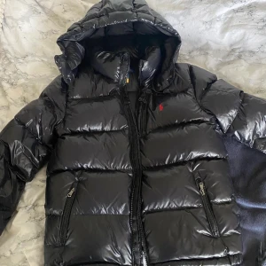 Svart pufferjacka från Polo Ralph Lauren - Säljer en svart pufferjacka från Polo Ralph Lauren med glansig finish och huva. Jackan har dragkedja framtill, två sidofickor med dragkedja och den klassiska röda loggan på bröstet. Perfekt för kalla dagar och riktigt snygg streetstil.