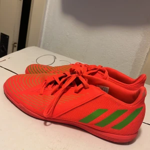 Adidas orange fotbollsskor med gröna detaljer - Snygga fotbollsskor från Adidas i en riktigt poppig orange färg med gröna ränder och mönster. Skorna har snörning och är gjorda i ett lätt syntetmaterial med meshpaneler för bra ventilation. Perfekta för dig som vill sticka ut på planen.