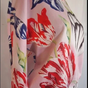 Blommig rosa tröja med lång ärm - Säljer en superfin tröja med stora blommor i rött, blått, svart och grönt på en ljusrosa bakgrund. Klänningen har lång ärm och rund halsringning, med ett färgstarkt och lekfullt mönster som verkligen sticker ut. Perfekt för dig som gillar att synas och vill ha något unikt i garderoben.