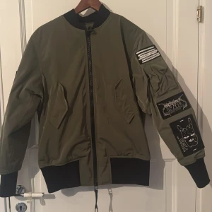 Olivgrön bomberjacka Tiger of Sweden - Nypris: 3700kr. Säljer en olivgrön bomberjacka från Tiger of Sweden Jeans med svarta muddar och coola svarta patches med vita tryck på ärm och rygg. Jackan har dragkedja framtill, två snedställda fickor och en riktigt streetig vibe. Perfekt för dig som vill sticka ut med din stil.