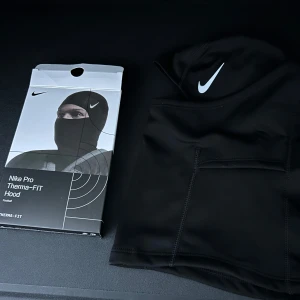 Nike Pro Therma-FIT svart balaclava - Nike Pro Therma-FIT balaclava i svart med vit swoosh-logga. Tight passform som täcker hela huvudet och halsen, perfekt för kalla dagar. Tillverkad i mjukt och stretchigt syntetmaterial som andas och håller värmen. Snygg och sportig look för träning eller utomhusaktiviteter.