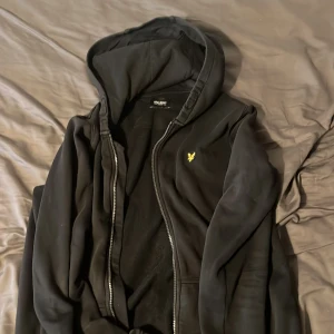 Svart hoodie från Lyle & Scott - Svart hoodie från Lyle & Scott med dragkedja och huva. Klassisk gul logga på bröstet och mjukt material som känns skönt mot huden. Perfekt för chill dagar eller när du vill ha en enkel och stilren look.