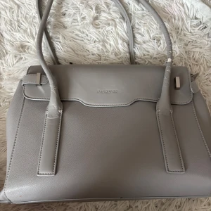 Grå handväska från Fiorelli - Snygg grå handväska från Fiorelli med stilren design och flera fack inuti för smart organisering. Väskan har två långa handtag och är tillverkad i slätt syntetmaterial. Perfekt för dig som gillar en clean och modern look. Ny köpt för 1200kr , repor i handtagen annars superfint skick 