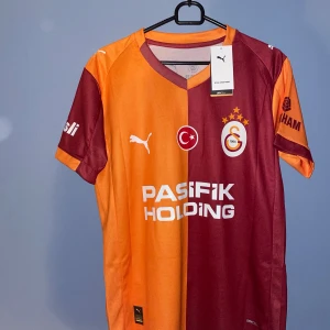 Galatasaray matchtröja Sané #10 Puma - Galatasaray fotbollströja från Puma med Sané och nummer 10 på ryggen. Tröjan har klassisk halvdelad design i vinrött och orange, korta ärmar och V-ringad krage. Materialet är lätt och ventilerande polyester med tryckta loggor och sponsorer. Perfekt för dig som älskar fotboll och vill sticka ut.