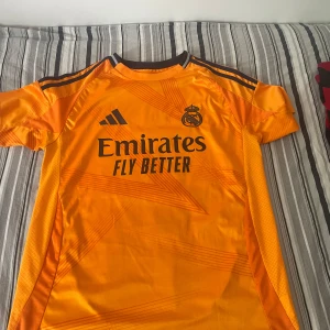 Real Madrid 24/25 borta kit Mbappé  - Snygg orange Real Madrid fotbollströja från Adidas med Mbappé och nummer 9 på ryggen. Tröjan har svarta detaljer, klubbmärke och sponsortryck framtill samt klassiska Adidas-ränder på axlarna. Tillverkad i lätt och ventilerande material, perfekt för fotboll eller träning. Ganska stor i storleken